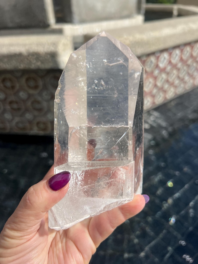 May include: A clear quartz crystal point with a faceted, geometric shape. The crystal is held in a hand with purple nail polish. The crystal is approximately 6 inches tall.