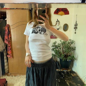 May include: A white short-sleeved t-shirt featuring a black graphic print of two figures embracing on the chest. The shirt is paired with a dark grey skirt that has an elastic waistband. The fabric of the t-shirt appears to be a ribbed knit material.