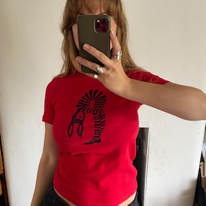 May include: A red t-shirt with a black graphic of a person bending backwards with a rabbit head. The short-sleeved shirt is made of a soft material.