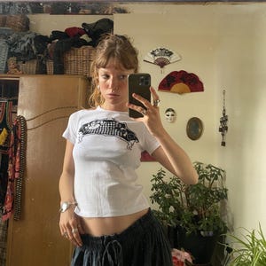 May include: A white short-sleeved t-shirt featuring a black graphic print on the chest. The graphic depicts a stylised, elongated shape with a chequered pattern and curved edges. The shirt has a crew neckline and a fitted, cropped silhouette.