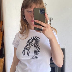 May include: A white short-sleeved t-shirt featuring a black and white graphic print of a figure with a spotted head and striped body bent backward in a flexible pose. The design is centred on the chest of the garment, which has a crew neckline.