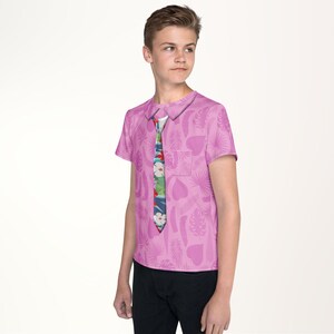 A young boy wearing a pink shirt with a tie pattern stands against a white background.