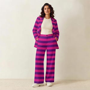 A woman wearing a purple and pink striped suit and white top stands in front of a white wall.