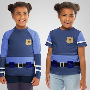 Puede incluir: Dos ni&ntilde;os con disfraces de polic&iacute;a azules. Los trajes presentan una insignia, un cintur&oacute;n con hebilla plateada y una franja azul claro en el pecho. Uno tiene mangas largas, el otro mangas cortas.