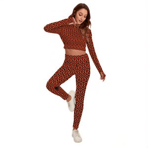 A woman wearing a red and black patterned outfit, including a crop top and leggings, is posing with one hand on her hip and the other hand on her thigh.