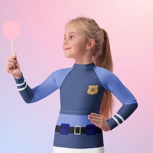 Puede incluir: Un disfraz de oficial de polic&iacute;a para ni&ntilde;os, con una parte superior de manga larga azul marino y azul claro con una insignia y detalles de cintur&oacute;n. El disfraz incluye una parte inferior blanca y se complementa con una piruleta rosa.