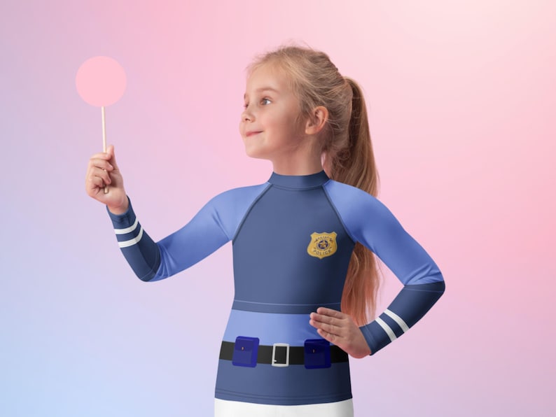 Puede incluir: Un disfraz de oficial de polic&iacute;a para ni&ntilde;os, con una parte superior de manga larga azul marino y azul claro con una insignia y detalles de cintur&oacute;n. El disfraz incluye una parte inferior blanca y se complementa con una piruleta rosa.