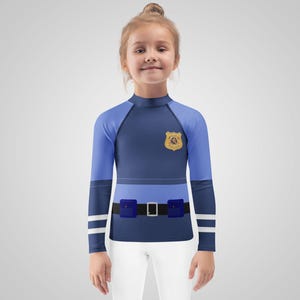 Puede incluir: Una camisa azul y azul claro de manga larga con un dise&ntilde;o de insignia de polic&iacute;a. La camisa presenta un cintur&oacute;n simulado con un cintur&oacute;n negro y una hebilla plateada, y pantalones blancos. La camisa est&aacute; dise&ntilde;ada para parecer un uniforme de polic&iacute;a.