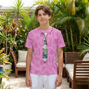 A young man wearing a pink shirt with a tie printed on it, standing in a tropical garden setting with palm trees and other plants in the background.