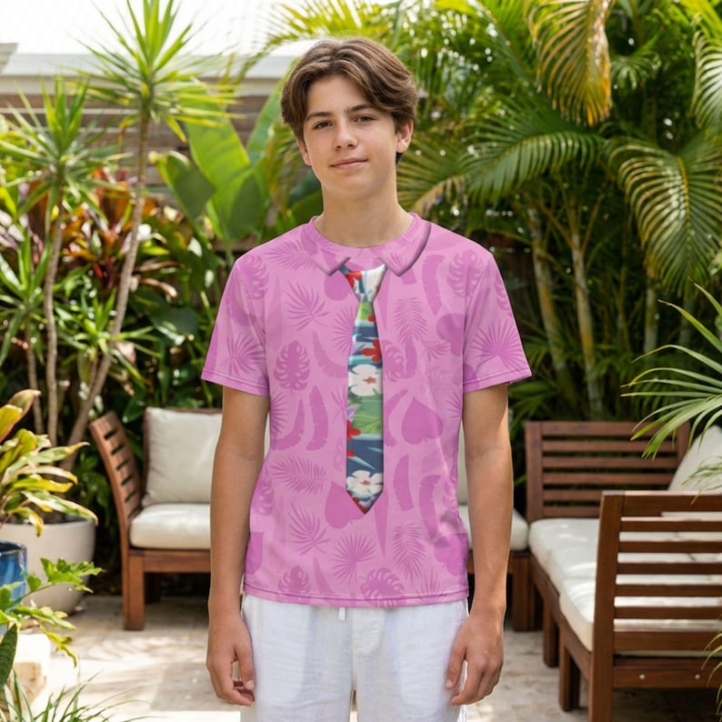 A young man wearing a pink shirt with a tie printed on it, standing in a tropical garden setting with palm trees and other plants in the background.
