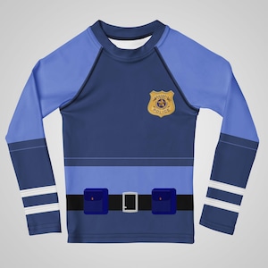 Puede incluir: Una camisa azul de manga larga dise&ntilde;ada para parecerse a un uniforme de polic&iacute;a. La camisa presenta una insignia, un cintur&oacute;n negro con hebilla plateada y dos bolsillos cuadrados azules. Las mangas tienen paneles azul claro y rayas blancas.