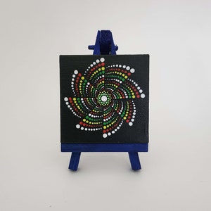May include: A small, square black canvas with a spiral dot design in white, red, yellow, and green, displayed on a miniature blue easel. The artwork features a central white dot, with the spiral expanding outwards.