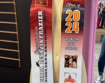 May include: A red and white graduation sash with the name "CIARA LEYAZIA FRAZIER" and the text "GOD IS WITHIN HER, SHE WILL NOT FALL, GOD WILL KEEP HER AT BREAK OF DAY PSALM 48:5". The sash also has a photo of a young girl and the text "Class of 2024 VIDALIA HIGH SCHOOL".