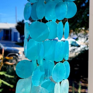 May include: A hanging wind chime with a wooden top and strands of translucent, light blue, shell-shaped pieces. The wind chime is suspended against a bright blue sky, suggesting an outdoor setting.