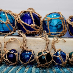 May include: A collection of glass fishing floats in shades of blue and teal, encased in woven rope netting. Some floats have a loop for hanging. The floats are arranged on a distressed white wooden surface, evoking a nautical theme.