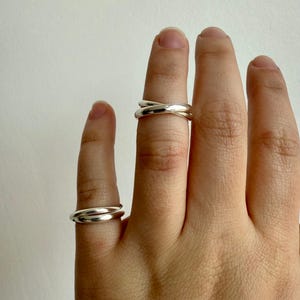 May include: Two silver rings are displayed on fingers against a neutral background. One ring has a simple band, while the other features an overlapping design. The rings are smooth and shiny, reflecting the light.