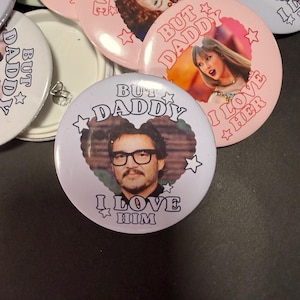 Op de afbeelding: Een verzameling ronde buttons met fotoportretten van verschillende beroemdheden. Elke button toont een hartvormig kader rond het portret, omringd door sterren en de tekst BUT DADDY I LOVE HIM of I LOVE HER.