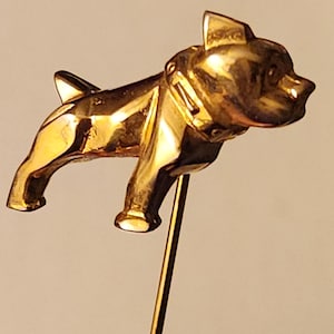 May include: A gold-coloured metal pin featuring a stylised bulldog design. The bulldog is standing on its hind legs with its front paws raised. The pin is attached to a thin metal rod.
