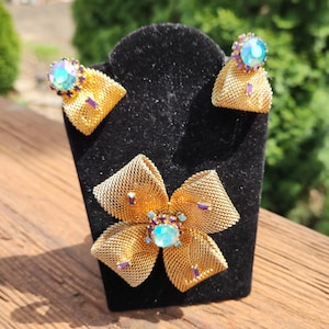 Puede incluir: Un broche de flor de malla dorada con piedras de cristal azul y morado iridiscentes. El broche se muestra con pendientes a juego.