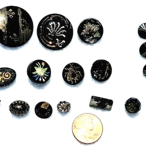 May include: A collection of black buttons in various shapes and sizes, featuring gold and colored painted designs, including floral, insect, and geometric patterns, displayed alongside a copper-colored penny for scale reference.