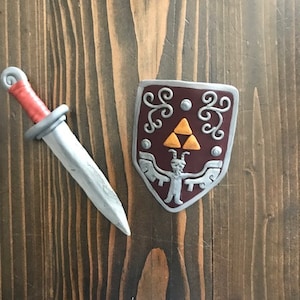 Op de afbeelding: Een zilveren zwaard met een rode handvat en een zilveren schild met een rode achtergrond en een gouden triforce-symbool. Het schild heeft een zilveren rand en een klein zilveren figuur met hoorns.