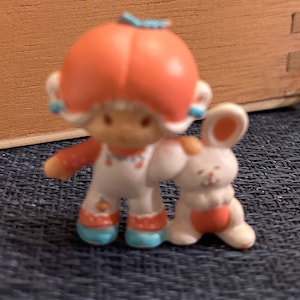 May include: A small orange and white figurine of a girl with a bunny rabbit. The girl has a large orange head with a blue bow and a white dress. The bunny is white with orange ears and a pink nose.