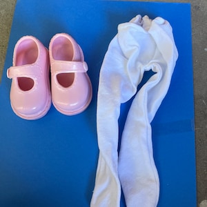 May include: A pair of pink doll shoes with a buckle and a pair of white doll tights.