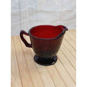 May include: A deep red glass creamer pitcher featuring a smooth, translucent finish, a curved handle on the side, a small pouring spout, and a circular pedestal base. The vessel has a wide, open top and a solid, dark ruby-colored glass construction.