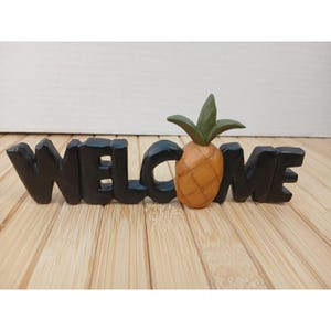 May include: A black carved sign spelling the word WELCOME in block letters, with the letter O replaced by a textured orange pineapple featuring green leaves. The sign has a matte finish and stands upright on a flat surface.
