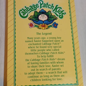 May include: A yellow box with a green and white Cabbage Patch Kids logo. The box has a yellow background with a white stripe pattern. The text on the box reads "Cabbage Patch Kids" and "The Legend". The text describes the origin of the Cabbage Patch Kids dolls.