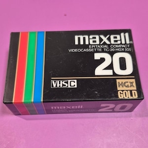 Könnte beinhalten: Eine schwarze Maxell VHS-C Videokassettenbox. Die Box zeigt den Text "maxell" in Weiß, zusammen mit "20" in großen weißen Zahlen. Ein vertikaler Streifen aus Rot, Grün, Blau und Gelb ziert die linke Seite. Die Box ist mit "HGX GOLD" beschriftet.
