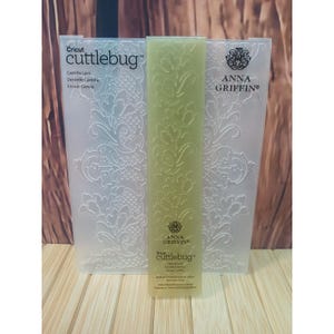 May include: A translucent plastic embossing folder by Cricut Cuttlebug and Anna Griffin titled Camilla Lace. The folder features a repeating floral and lattice pattern. The packaging includes the brand names and the product title in white text.
