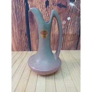 May include: A ceramic pitcher with a matte finish in muted mauve and sage green tones. The vessel features a tall, slender neck, a wide base, and a curved handle. A gold-colored label on the front reads NILOAK POTTERIES BENTON ARKANSAS.