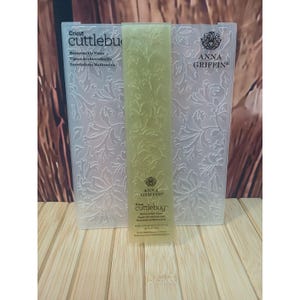 May include: A translucent plastic embossing folder by Cricut Cuttlebug and Anna Griffin featuring a honeysuckle vine pattern. The rectangular folder is embossed with floral designs and includes the text Cricut Cuttlebug and Anna Griffin.