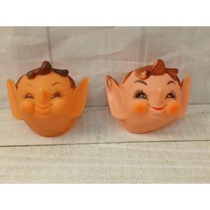 May include: Two molded rubber elf heads with pointed ears, brown hair, and painted facial features. The left elf is orange-toned, while the right elf is a lighter peach color. Both figures feature wide smiles, rosy cheeks, and long eyelashes.