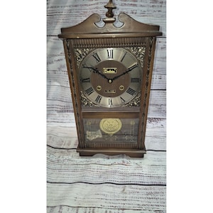 May include: A brown wooden wall clock with a decorative top finial. The clock face features a silver-toned dial with black Roman numerals, black hands, and the text Centurion and 35 DAY. A gold-toned pendulum is visible behind a glass panel.