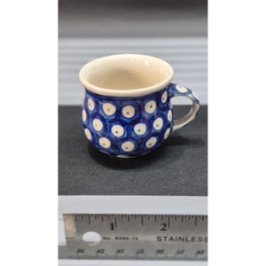 May include: A small, ceramic mug with a blue background and white polka dots. The mug has a cream-colored interior and handle. The mug is approximately 2 inches tall.