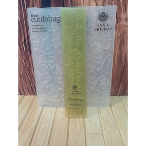 May include: A translucent, rectangular embossing folder by Cricut Cuttlebug and Anna Griffin titled Scripted Love. The folder features a repeating embossed script pattern of the word Love and a central vertical column of stylized leaf motifs.