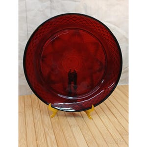 May include: A circular ruby red glass plate featuring a diamond-patterned rim and a smooth, clear center. The plate is displayed on a small, gold-colored metal stand, showcasing its translucent deep red hue and reflective surface texture.