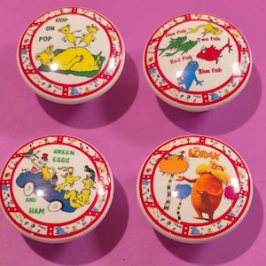 May include: Four white ceramic cabinet knobs with colorful illustrations from Dr. Seuss books. The knobs feature images and text from "Hop on Pop", "One Fish Two Fish Red Fish Blue Fish", "Green Eggs and Ham", and "The Lorax". Each knob has a red border.