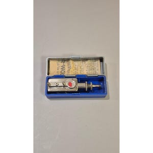 May include: A vintage metal tool kit with a blue plastic interior. The kit contains a silver-colored tool with a red button and a threaded component. A small piece of paper with text is also inside the kit.