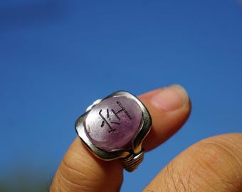 Puede incluir: Un anillo hecho a mano con una piedra ovalada de color púrpura claro grabada con las iniciales "KH" en negro. La piedra está engastada en una banda plateada envuelta en alambre. El anillo se muestra contra un cielo azul brillante.
