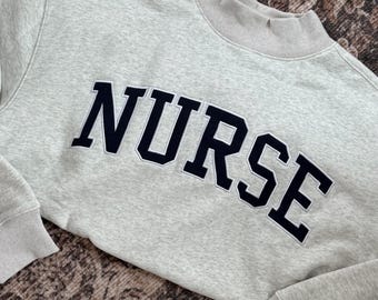 May include: A light gray heathered sweatshirt featuring a mock neck collar and ribbed cuffs. The word NURSE is embroidered across the chest in bold, dark navy blue capital letters with a thin white outline, creating a collegiate style.