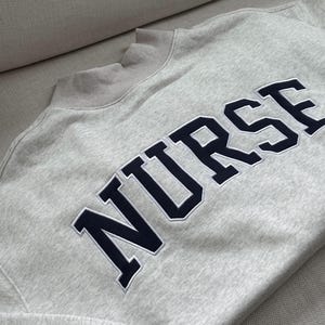 May include: A light grey sweatshirt featuring the word "NURSE" in navy blue, collegiate-style lettering. The sweatshirt has a crew neck and ribbed cuffs and hem.
