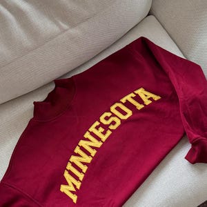 May include: A maroon long-sleeve sweatshirt featuring the word MINNESOTA in large, bold, yellow embroidered capital letters across the chest. The garment has a mock neck collar and ribbed cuffs, showcasing a classic athletic design style.