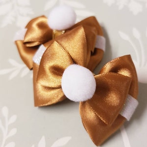 Puede incluir: Dos lazos para el pelo de satén dorado con pompones blancos y detalles de purpurina blanca. Los lazos están atados con forma de lazo clásico, probablemente para un niño o mascota. Sobre un fondo verde claro.