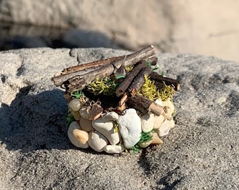 Può includere: Una casa in miniatura di pietra e ramoscelli con muschio verde sul tetto, seduta su una superficie grigia e sabbiosa.