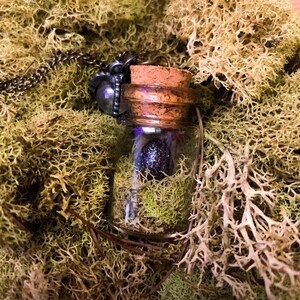 May include: A glass bottle necklace with a cork stopper and a dark purple, glittery orb inside. The bottle is filled with green moss and is hanging from a silver chain.