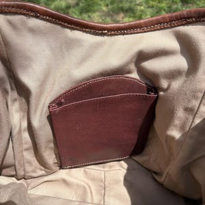 May include: Inside view of a brown leather handbag with a burgundy leather pocket. The bag's interior is a light tan fabric, and the top edge is trimmed with brown leather. A gold zip is visible at the bottom.