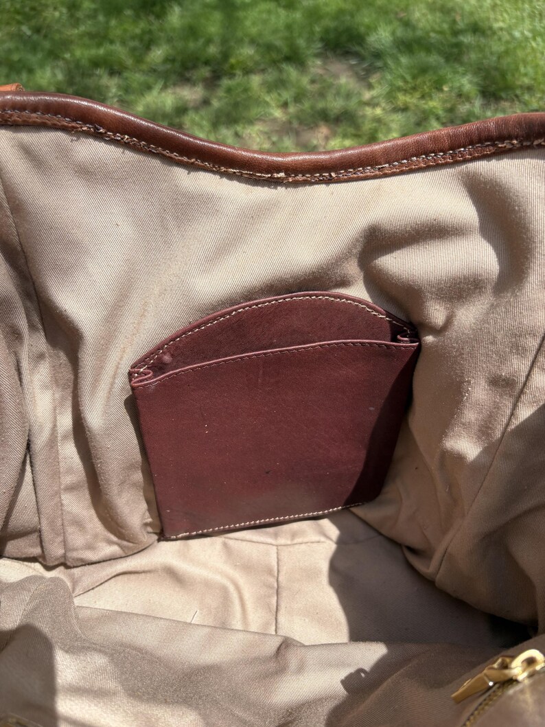 May include: Inside view of a brown leather handbag with a burgundy leather pocket. The bag's interior is a light tan fabric, and the top edge is trimmed with brown leather. A gold zip is visible at the bottom.